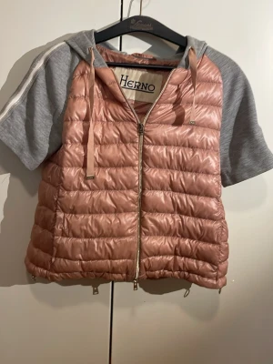Rosa dunväst med huva från Herno - Supersnygg rosa dunväst från Herno med quiltad design och dragkedja framtill. Västen har en grå huva och gråa korta ärmar i sweatshirtmaterial samt dragsko i huvan. Perfekt för lager på lager och streetstyle. Inte så använd. Inga hål.