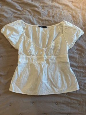 Vit blus från Brandy Melville - Den har en liten fläck på armen men det syns knappast och kan nog också fixas rätt lätt. Söt vit blus från Brandy Melville med korta ärmar och djup urringning. Blusen har knappar framtill och lätt rynkade detaljer vid bysten. Tillverkad i ett tunt, luftigt bomullstyg som ger en avslappnad och somrig känsla.