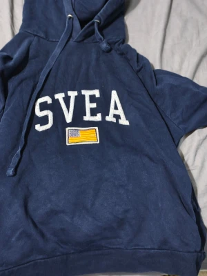 Mörkblå hoodie från SVEA - Mörkblå hoodie från SVEA med stor vit logga på bröstet och en broderad gul och blå flagga under. Klassisk huva med dragsko och mjukt material i bomullsblandning. Perfekt för en avslappnad och sportig stil.