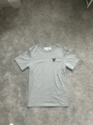 Grå Comme des Garçons PLAY t-shirt - Snygg grå t-shirt från Comme des Garçons PLAY med det ikoniska svarta hjärta-trycket på bröstet. Klassisk passform med rund hals och korta ärmar. Perfekt till jeans eller shorts för en clean och stilren look.