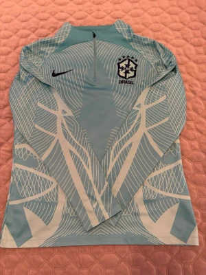 Nike Brasil träningströja ljusblå - Nike träningströja för Brasilien i ljusblått med vitt grafiskt mönster. Halv dragkedja framtill och lång ärm. Perfekt för fotbollsträning eller uppvärmning. Brasil-logga och Nike Swoosh på bröstet.