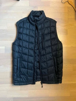 Svart quiltad dunväst - Snygg svart dunväst med quiltat mönster och hög krage. Västen har dragkedja framtill, två sidofickor och en bröstficka med dragkedja. Perfekt att slänga över en hoodie eller tröja för extra värme. Klassisk och stilren design.