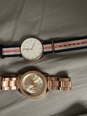 Två stilrena klockor DW & Michael Kors - Säljer två snygga klockor! En Daniel Wellington med vit urtavla och randigt blått, vitt och rosa tygarmband. En Michael Kors i roséguld med metallänk och glittrande stenar runt. Säljer båda tillsammans men kan även sälja dom separat.