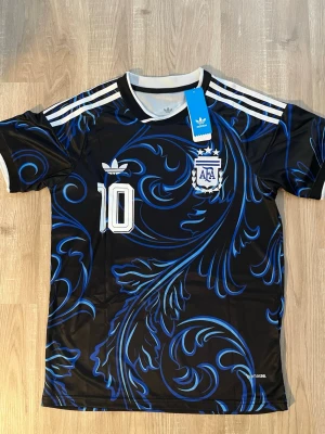 Argentina Messi VM-2026 Fotbollströja - Säljer en svart och blå Argentina fotbollströja med Messi och nummer 10 på ryggen. Tröjan har vita detaljer, AFA-logga och Adidas ränder på axlarna. Tillverkad i ett lätt och ventilerande material, perfekt för träning eller match. HELT NY