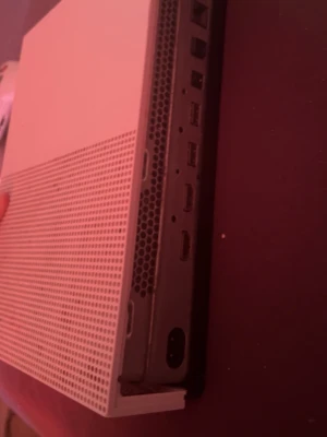 Xbox One S vit spelkonsol med handkontroll - Säljer en Xbox One S i snygg vit design med tillhörande vit trådlös handkontroll. Konsolen har en stilren, rektangulär form med ventilerade sidor och flera anslutningsportar på baksidan. Perfekt för gaming och streaming i vardagsrummet.
