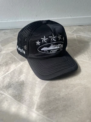 Svart Crtz keps med mesh och brodyr - Säljer en svart keps från Crtz med mesh på sidorna och broderade stjärnor samt ett motiv framtill. Justerbar snapback baktill och vit brodyr på baksidan. Perfekt för dig som vill ha en stilren och streetinspirerad look.
