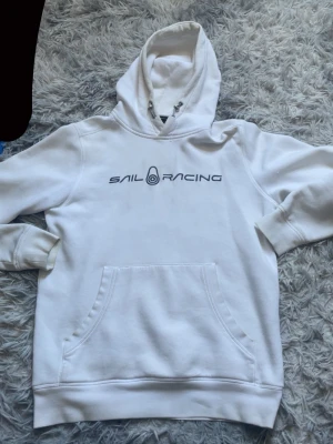 Vit hoodie från Sail Racing - Säljer en vit hoodie från Sail Racing med stor logga tryckt på bröstet. Hoodien har en klassisk huva med dragsko och en stor magficka framtill. Tillverkad i mjukt bomullsmaterial som är skönt mot huden. Perfekt för en avslappnad och sportig stil.