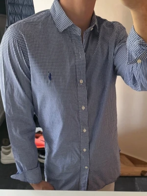 Blårutig skjorta från Ralph Lauren - Klassisk blårutig skjorta från Ralph Lauren med vit knappstängning och broderad logga på bröstet. Skjortan har lång ärm, krage och är tillverkad i bomull. Perfekt för en stilren look. Storlek 38 vilket motsvarar m typ