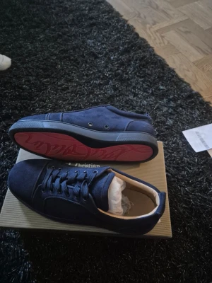 Marinblå Louboutin sneakers - Snygga marinblå sneakers från Christian Louboutin i mocka med klassisk röd sula och diskret logga på hälen. Skorna har lågt skaft, svarta snören och en stilren design som passar till det mesta. Perfekta för dig som vill ha något exklusivt och trendigt.