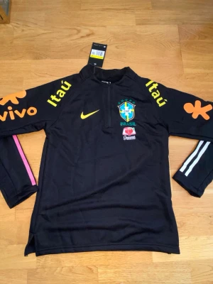 Svart Brasilien Nike träningströja - Svart långärmad träningströja från Nike med Brasilien CBF-logga, gula och orangea sponsortryck samt halv dragkedja. Tröjan har detaljer i rosa och vitt på ärmarna och är tillverkad i funktionsmaterial. Perfekt för fotbollsträning eller sportiga aktiviteter.