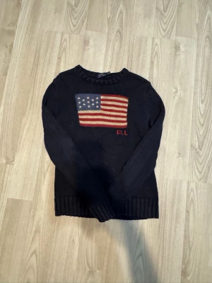 Mörkblå stickad tröja Polo Ralph Lauren - Mörkblå stickad tröja från Polo Ralph Lauren med amerikansk flagga i rött, vitt och blått på bröstet samt RL-broderi. Tröjan har rund halsringning och ribbstickade muddar vid ärmslut och nederkant. Perfekt för dig som gillar klassisk preppy stil.