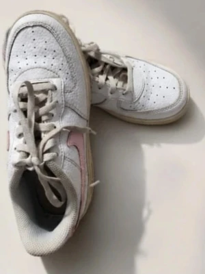 Nike Air Force 1 vita & rosa sneakers - Nike Air Force 1 sneakers i vitt läder med ljusrosa swoosh och detaljer. Klassisk låg modell med perforerad tå, vita snören och beige sula. Perfekta för dig som vill ha en clean men ändå unik look. Passar till jeans, kjol eller shorts.