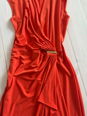 Orange omlottklänning Michael Kors - Snygg orange omlottklänning från Michael Kors med draperad effekt och guldfärgad detalj i midjan. Klänningen är ärmlös och har en mjuk, följsam känsla i tyget som ger en elegant siluett. Perfekt för dig som vill sticka ut med färg och stil. Nypris 2500kr