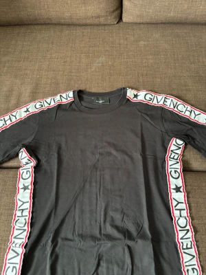 Svart Givenchy t-shirt med logoband - Svart t-shirt från Givenchy med vita och röda logoband längs axlar och sidor där det står 'GIVENCHY' och stjärnor. Klassisk rund halsringning och korta ärmar. Perfekt för dig som gillar streetwear och exklusiva märken.