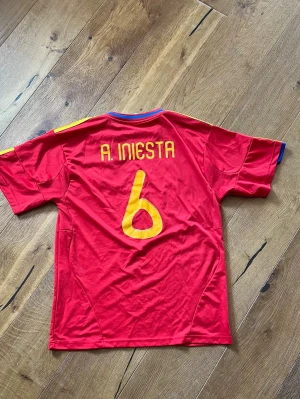 Spansk landslagströja Iniesta #6 - Röd matchtröja från Spaniens landslag med Iniesta och nummer 6 i gult på ryggen. Broderat landslagsemblem med stjärna på bröstet. Klassisk fotbollströja i funktionsmaterial, perfekt för träning eller match.