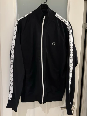 Svart track jacket från Fred Perry - Snygg svart track jacket från Fred Perry med vita detaljer och dragkedja framtill. Klassiska vita ränder med Fred Perry-logga längs ärmarna och broderad logga på bröstet. Hög krage och ribbade muddar. Perfekt för en sportig och trendig look.
