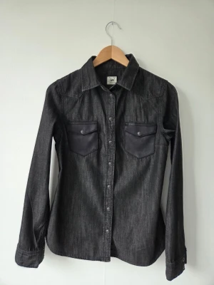 Mörkgrå jeansskjorta från Lee - Snygg mörkgrå jeansskjorta från Lee med klassisk krage och två bröstfickor med knappar. Skjortan har långa ärmar och tryckknappar framtill. Perfekt för en cool och avslappnad stil.