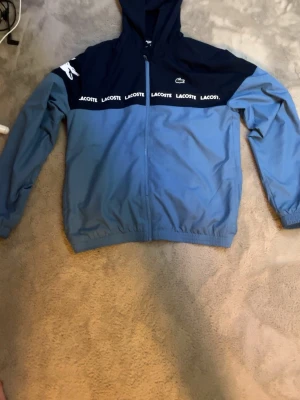 Blå vindjacka med huva från Lacoste - Snygg tvåfärgad vindjacka från Lacoste i ljusblått och marinblått. Jackan har huva, dragkedja framtill och elastiska muddar. Dekorativ Lacoste-logga och text på bröstet samt en stor krokodil på axeln. Perfekt för en sportig och trendig look.