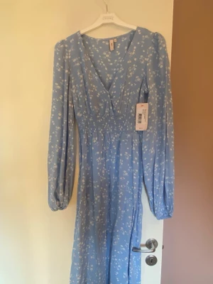 Blå blommig långklänning Nelly - Söt långklänning i ljusblått från Nelly  med små vita blommor. Klänningen har långa puffärmar, v-ringning och smock i midjan för en snygg siluett. Tillverkad i ett lätt, flowy tyg som ger en romantisk känsla.