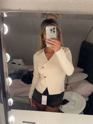 Krämvit kofta med guldknappar - Snygg krämvit kofta från ZARA med figurnära passform och dekorativa guldknappar framtill. Koftan har V-ringning och långa ärmar, perfekt att styla till både jeans och kjol. Materialet känns mjukt och stretchigt. Aldrig använd då de inte riktigt va min stil. 