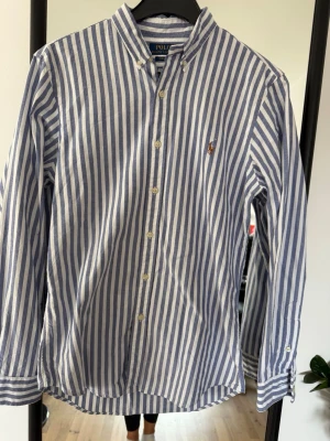 Randig skjorta från Polo Ralph Lauren - Klassisk skjorta från Polo Ralph Lauren med blå och vita vertikala ränder. Skjortan har lång ärm, knappar framtill och den ikoniska broderade loggan på bröstet. Tillverkad i mjuk bomull och har en tidlös, normal passform.