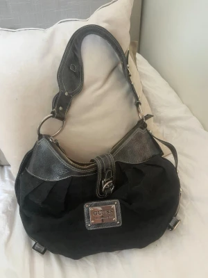 Svart axelremsväska från Guess - Sjukt snygg svart axelremsväska från Guess med detaljer i konstläder och metall. Väskan har ett stort fack med dragkedja, leopardmönstrat innerfoder och en metallplatta med Guess-logga framtill. Perfekt för dig som gillar statement-accessoarer. Det är en äldre modell och den har använts, men den är fortfarande i fint skick med bara bl.a. några slitningar på spännena. 