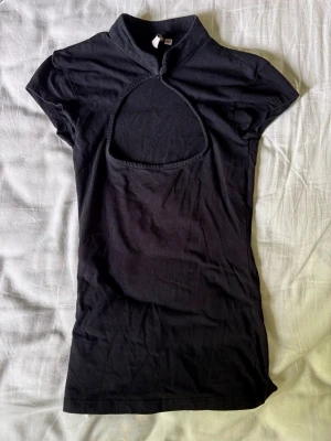Svart topp med cut-out detalj - Trendig svart topp med hög krage och unik cut-out detalj framtill. Kortärmad och figurnära passform, perfekt för att skapa en edgy look. Tillverkad i mjukt bomullsmaterial som känns bekvämt mot huden.