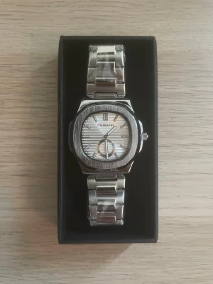 Silverfärgad klocka från MAIMASHI  - Stilren klocka från MAIMASHI i silverfärg med metallarmband och fyrkantigt ur. Urtavlan är vit med diskreta ränder och har en extra liten sekundvisare. Perfekt accessoar för dig som gillar en clean och modern look.