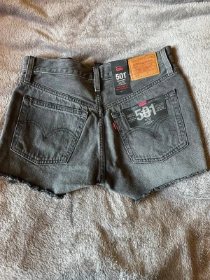 Grå Levi's 501 jeansshorts - Säljer ett par grå Levi's 501 jeansshorts med råa, fransiga kanter och klassisk femficksdesign. Har aldrig används och har prislappen kvar. Shortsen har hög midja och är tillverkade i bomull. Perfekta för en avslappnad och cool stil.