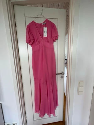 Hot pink maxiklänning från Adoore - Säljer en snygg hot pink maxiklänning från Adoore med v-ringning och korta puffärmar. Klänningen har en figurnära passform upptill och faller snyggt nedtill med ett lätt, flowy tyg. Perfekt för dig som vill sticka ut med färg och stil.