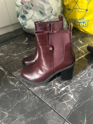 Vinröda stövletter med klack - Snygga vinröda stövletter med bred klack och dragkedja på sidan. Tillverkade i ett glansigt skinnliknande material med elastisk panel och dekorativ knappdetalj. Perfekta för dig som vill ha en stilren och trendig look.