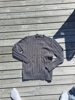 Grå stickad tröja Polo Ralph Lauren - Snygg grå stickad tröja från Polo Ralph Lauren med klassiskt kabelstickat mönster. Tröjan har rund halsringning och ribbade muddar vid ärmslut och nederkant. Perfekt för dig som gillar stilrena och tidlösa plagg.