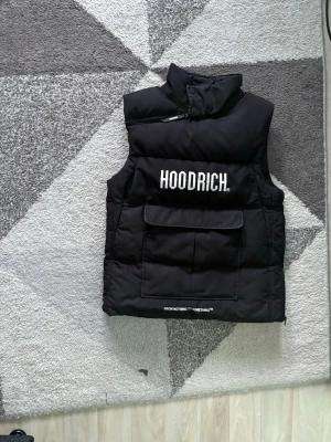 Svart dunväst från Hoodrich - Snygg svart dunväst från Hoodrich med hög krage och stor ficka framtill. Västen har tydlig vit logga på bröstet och textdetalj längst ner. Perfekt för lager-på-lager och streetwear-stil.