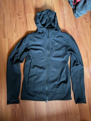 Svart Nike hoodie med dragkedja - Snygg svart hoodie från Nike med hel dragkedja framtill och diskret Nike-logga på bröstet. Hoodien har en åtsittande passform, långa ärmar och praktisk huva. Tillverkad i mjukt och stretchigt material som passar perfekt till en avslappnad stil.