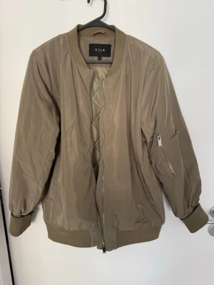Beige bomberjacka från Vila - Bomberjacka från Vila med quiltad insida. Jackan har dragkedja framtill och en ficka med dragkedja på ärmen. Perfekt för dig som vill ha en enkel men snygg jacka till vardags.