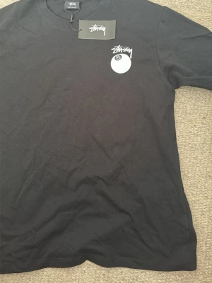 Svart Stüssy t-shirt med 8-ball print - Svart t-shirt från Stüssy med ikoniskt 8-ball tryck och Stüssy-logga både fram och bak. Klassisk rund hals och korta ärmar. Tillverkad i mjuk bomull för skön passform. Perfekt för dig som gillar streetwear och vill sticka ut.