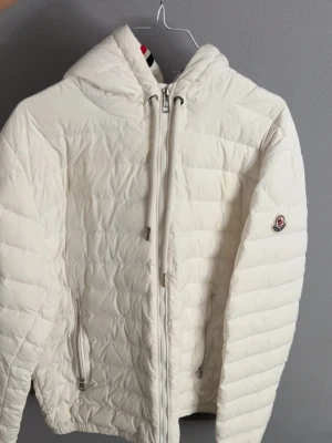Vit quiltad dunjacka från Moncler - Snygg vit dunjacka från Moncler med quiltad design och huva. Jackan har dragkedja framtill, två sidofickor med dragkedja och Moncler-logga på ärmen. Huvan har coola ränder i rött, blått och vitt. Perfekt för kalla dagar! SE SISTA BILD FÖR LITEN DEFEKT