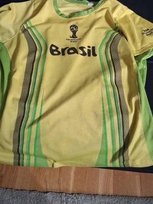 Gul Brasil fotbollströja VM 2014 - Snygg gul Brasilien fotbollströja från VM 2014 med gröna och svarta detaljer, tryck på bröstet och ryggen samt nummer 10. Tillverkad i lätt och ventilerande material, perfekt för träning eller match. Klassisk sportig design med korta ärmar.En retro tröja som är rare.