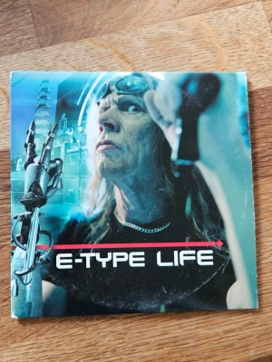 E-Type Life CD-singel - CD-singel med E-Type, låten 'Life' feat. Na Na. Omslaget har futuristiskt tema med robotarm och blåa toner. Baksidan listar två versioner av låten och har dekorativa mönster i hörnen. Perfekt för dig som gillar eurodance och samlar på 90-talsmusik.