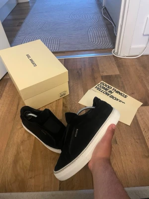 Svarta sneakers från Axel Arigato - Stilrena svarta sneakers från Axel Arigato med vit sula och minimalistisk design. Skorna är låga och har en ovandel i mocka med diskret logga på sidan. Perfekta för dig som gillar en clean och modern look.
