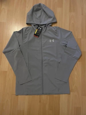 Under Armour Windbreaker  - Snygg grå Windbreaker från Under Armour med dragkedja framtill och huva. Tillverkad i ett mjukt och stretchigt material som passar perfekt till träning eller chill. Under Armour-logga på bröstet och stort tryck på ryggen. Långärmad och sportig design.TVEKA INTE ATT SKICKA EN BUD:)