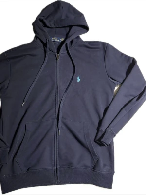 Marinblå hoodie från Polo Ralph Lauren - Snygg marinblå hoodie från Polo Ralph Lauren med dragkedja, huva med dragsko och två stora fickor framtill. Klassisk logga broderad på bröstet i ljusblått. Tillverkad i mjuk bomullsblandning och har ribbade muddar vid ärmslut och nederkant.