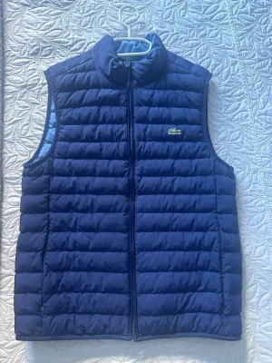 Mörkblå dunväst från Lacoste - Snygg mörkblå dunväst från Lacoste med klassisk quiltad design och dragkedja framtill. Västen har det ikoniska gröna krokodilmärket på bröstet och står för både stil och komfort. Perfekt att slänga över en hoodie eller tröja när det är kyligt ute.