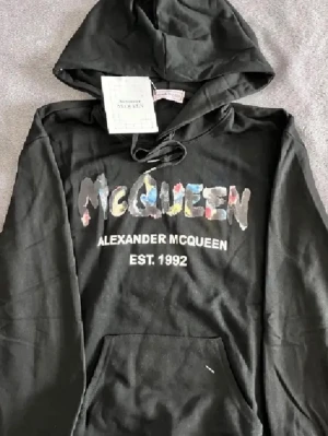 Svart hoodie Alexander McQueen - Snygg svart hoodie från Alexander McQueen med färgglad logga framtill och texten 'EST. 1992'. Klassisk känguruficka och justerbar huva med snören. Perfekt för dig som vill ha en streetwear vibe och sticka ut lite extra. Mjuk bomullskänsla.