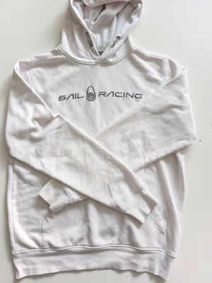 Vit hoodie från Sail Racing Storlek M  - Säljer en vit hoodie från Sail Racing med tryckt logga på bröstet. Hoodien har huva med dragsko och känguruficka framtill. Tillverkad i mjukt bomullsmaterial som känns skönt mot huden. Perfekt för en avslappnad och sportig stil.