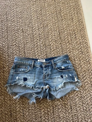 Blå slitna jeansshorts med fransar - Säljer ett par blå jeansshorts från One Teaspoon med råa, fransiga kanter och slitna detaljer framtill. Shortsen har klassisk femficksmodell, knappgylf och låg midja. Perfekta för sommaren och ger en avslappnad vibe.