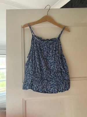 Blommig blå linnetopp från H&M - Söt blå linnetopp från H&M med små vita blommor och tunna axelband. Toppen har en avslappnad passform och är perfekt för varma dagar. Tillverkad i lätt och luftig polyester som känns skön mot huden.