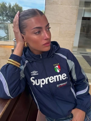 Mörkblå Supreme x Umbro vindjacka - Snygg mörkblå vindjacka från Supreme och Umbro med vita detaljer och dragkedja. Jackan har stor Supreme-logga framtill, Umbro-märke och broderad sköld med stjärnor. Lätt material, huva och sportig look. Perfekt för streetwear.