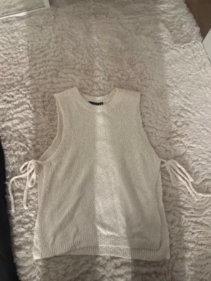 Stickad vit väst med snörning i sidan - Mysig stickad väst i vit färg med rund halsringning och snörningsdetaljer på båda sidor. Perfekt att bära över en skjorta eller t-shirt för en trendig lager-på-lager-look. Västen har en avslappnad passform och ribbade kanter.