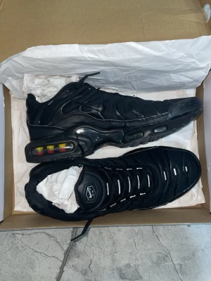 Nike Air Max Plus svarta sneakers - Nike Air Max Plus sneakers i helsvart med mesh och syntetiska detaljer. Skorna har synlig Air Max-dämpning med gula och röda inslag i sulan, samt reflekterande detaljer på ovandelen. Ikonisk TN-logga på plösen och en sportig, chunky siluett.